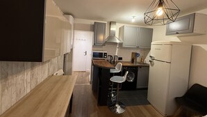 Cocina compacta con encimera de madera, microondas integrado, fregadero y refrigerador. Hay un taburete de bar y una lámpara colgante sobre la encimera.