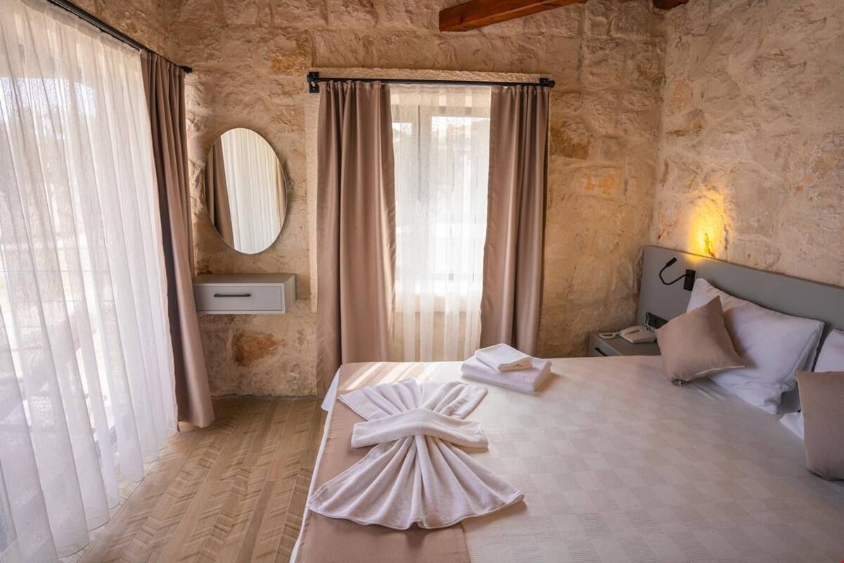 Habitación de hotel con pared de piedra, cama con toallas ordenadas, teléfono en la mesita de noche y ventana con cortinas translúcidas.