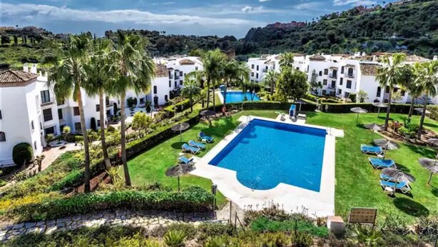 A resort with a central swimming pool surrounded by white buildings, palm trees, and a hilly landscape in the background. - 2 Bedrooms - Sleeps 4 - Garden and Pool With View (Benahavis)