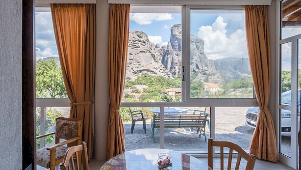 A room with a large window offering a view of rocky mountains and a distant village. Inside, there is a round table with chairs and a small decorative item on the table. - Majestic view Home just under Meteora Unesco Site

 (PJ57+9V6 Kalabaka)