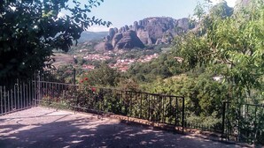 A paved pathway with a metal fence and flowering plants, leading to a town with red-roofed buildings and rocky cliffs in the background. - Majestic view Home just under Meteora Unesco Site

 (PJ57+9V6 Kalabaka)