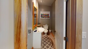 A narrow hallway leading to a bathroom with a white sink, toilet, and a mirror. A colorful artwork is visible on the wall above the toilet.