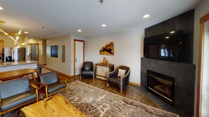 A modern living room with a fireplace, a flat-screen TV, and a dining area with a kitchen in the background.