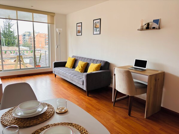 A modern living room with a grey sofa, yellow cushions, a wooden desk with a laptop, and a dining table set with plates and glasses.
