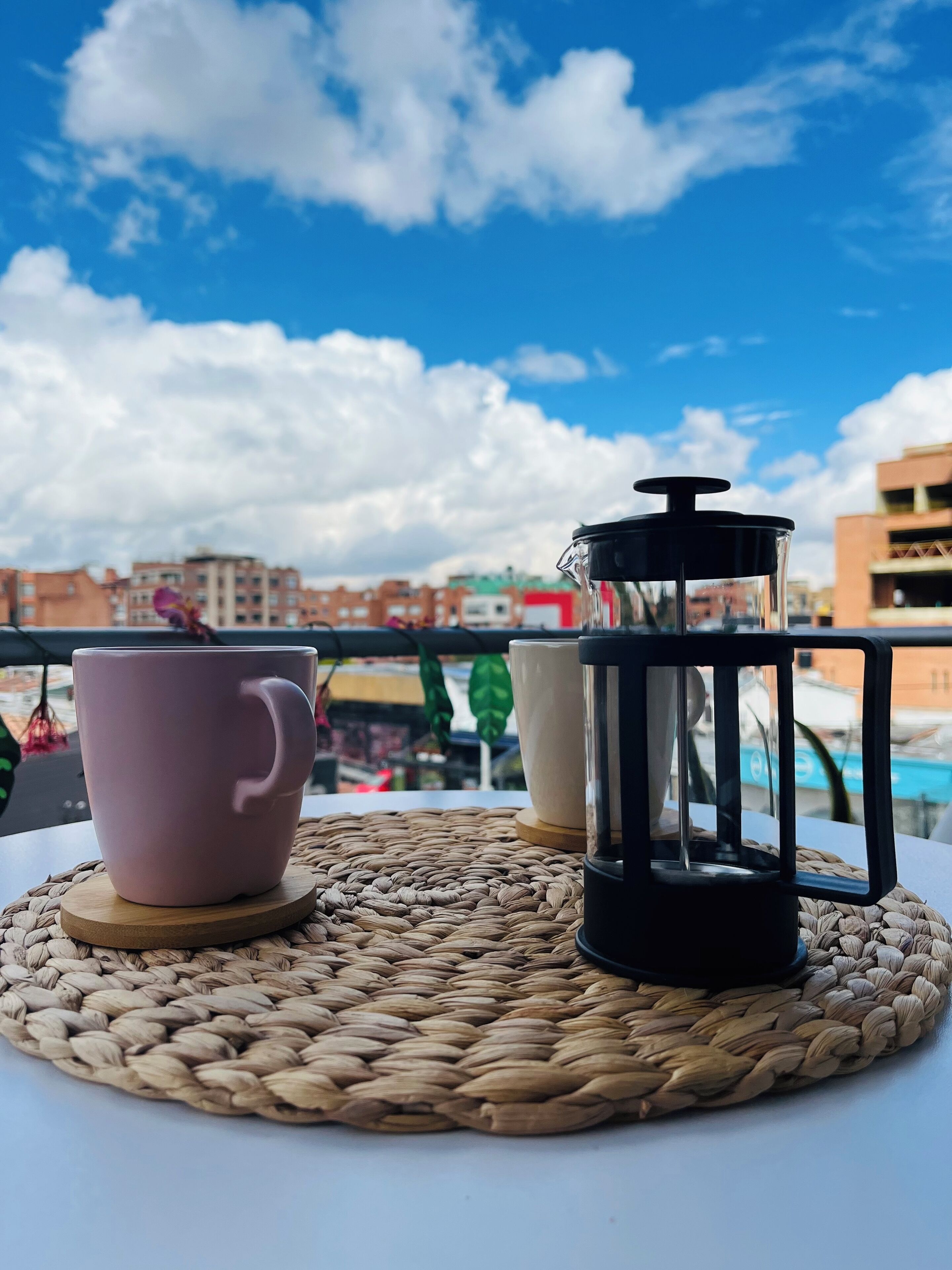 Uma mesa com um descanso trançado, uma caneca rosa e uma cafeteira preta em uma varanda com uma vista urbana ao fundo.