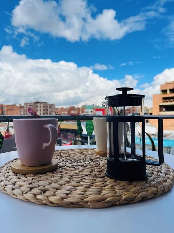 A table with a woven placemat, a pink mug, and a black coffee pot on a balcony with a cityscape in the background.