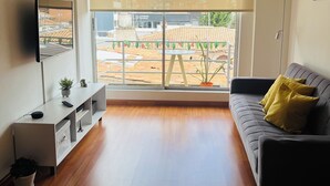 Een moderne woonkamer met een groot raam, een grijze bank met gele kussens, een witte tv-meubel en een vloerlamp.