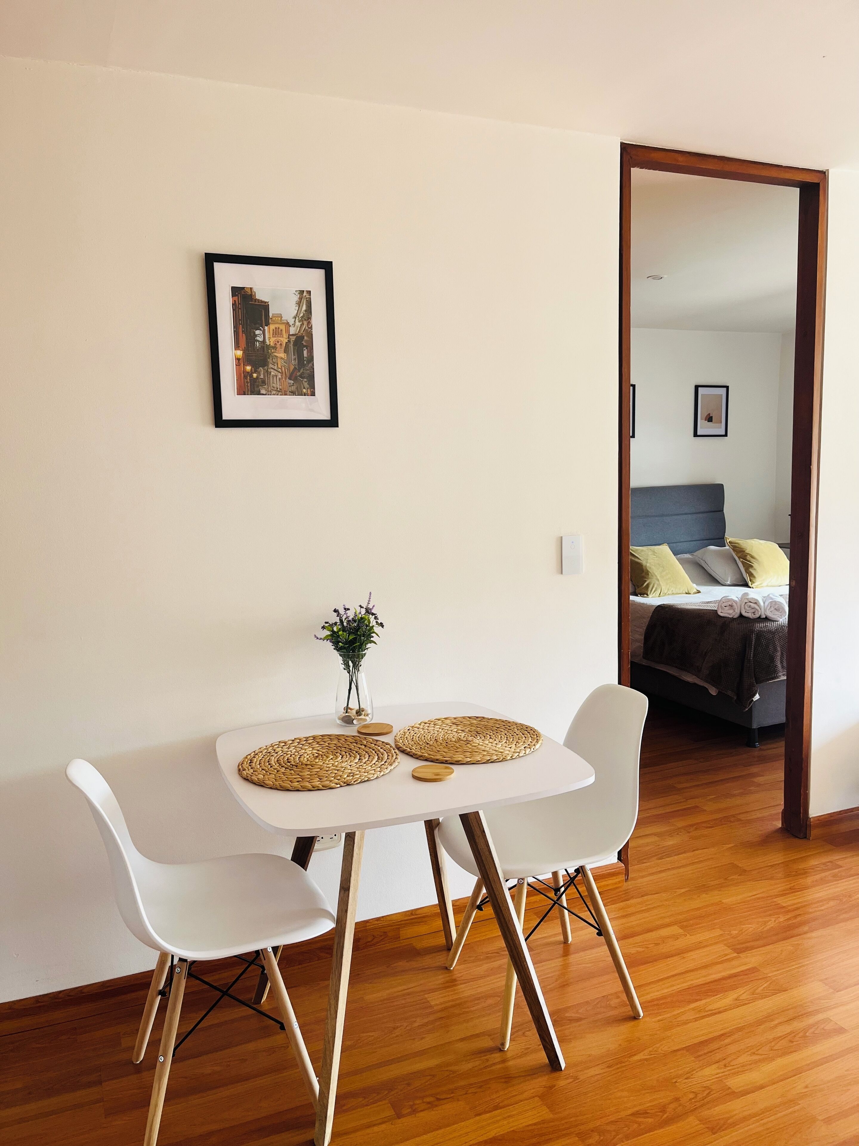 A room with a white table, two white chairs, a small vase with flowers, and a framed picture on the wall.
