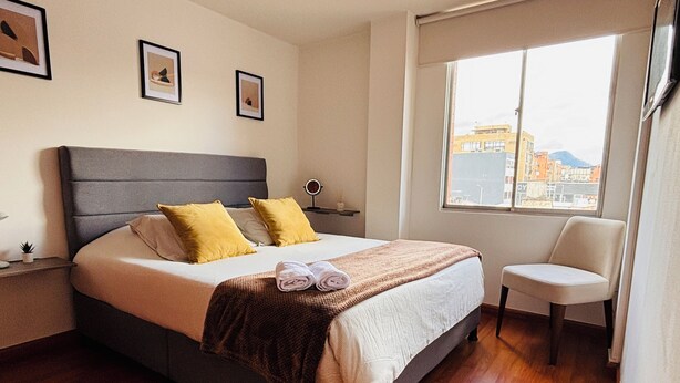 Een netjes opgemaakt bed met een grijs hoofdbord, twee gele kussens en een bruine plaid. Er staat een klein nachtkastje met een lamp bij het bed, en aan de muur hangt een ingelijste foto. Een stoel staat naast het bed, en een raam biedt uitzicht op gebouwen.