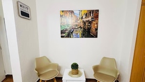 A room with a white wall, a painting of a canal, two beige chairs, a small white table with a plant, and a WiFi password sign.