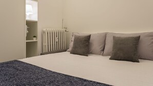 Une chambre à coucher avec un lit, des oreillers, un radiateur et une étagère sur laquelle se trouvent une lampe et une plante.
