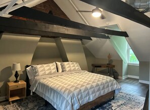 A bedroom with a sloped ceiling, a bed with a patterned bedspread, a nightstand with a lamp, and a small table with a lamp.