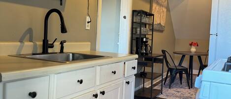 A kitchen with white cabinets, a black faucet, and a patterned rug. There is a dining area with a table and chairs, and a refrigerator in the corner.