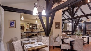 A restaurant with high ceilings, exposed beams, and patterned upholstered chairs. Tables are set with white tablecloths and silverware.