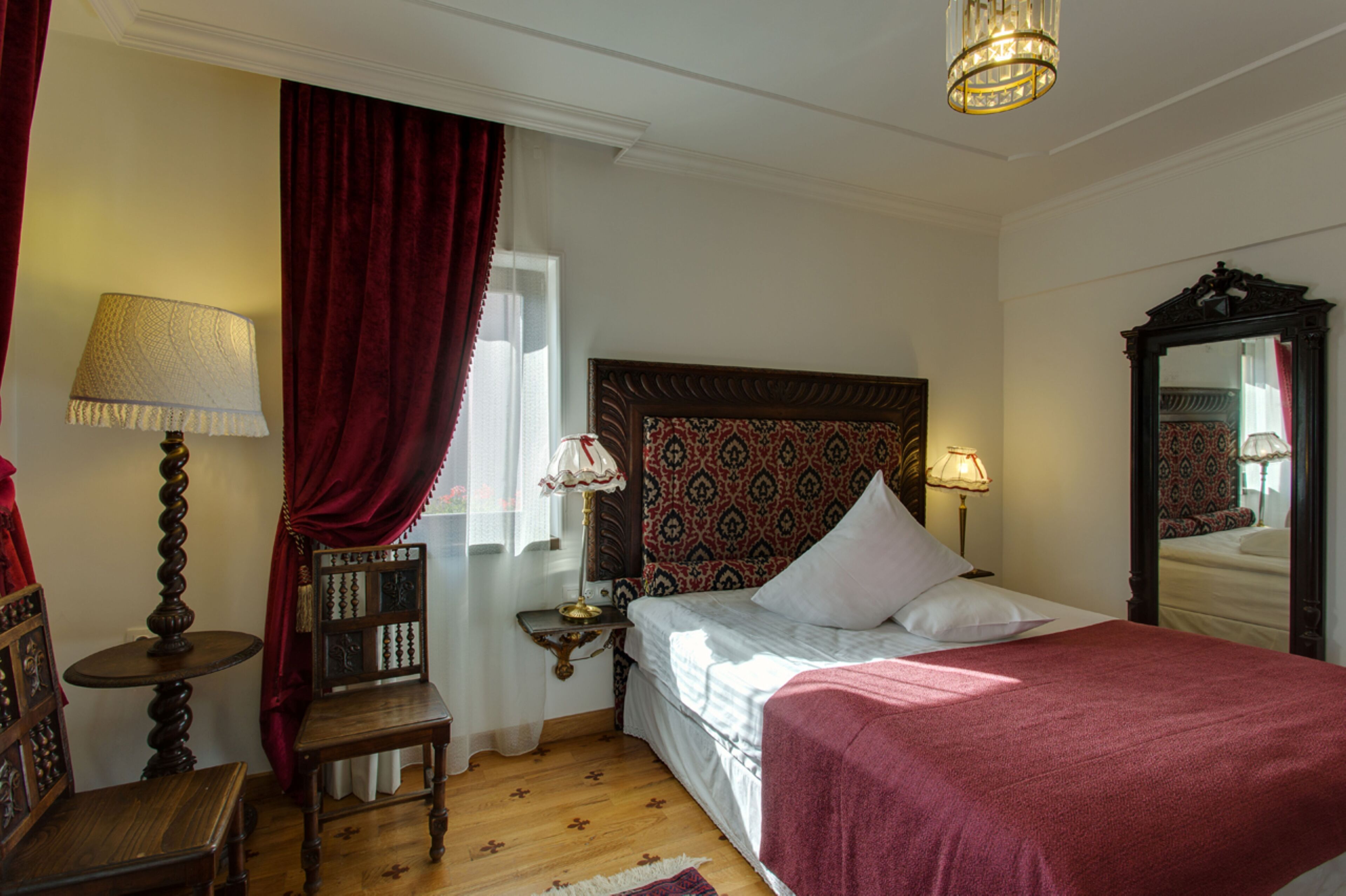 A hotel room with a bed, a nightstand, a chair, a lamp, a mirror, and a window with curtains.