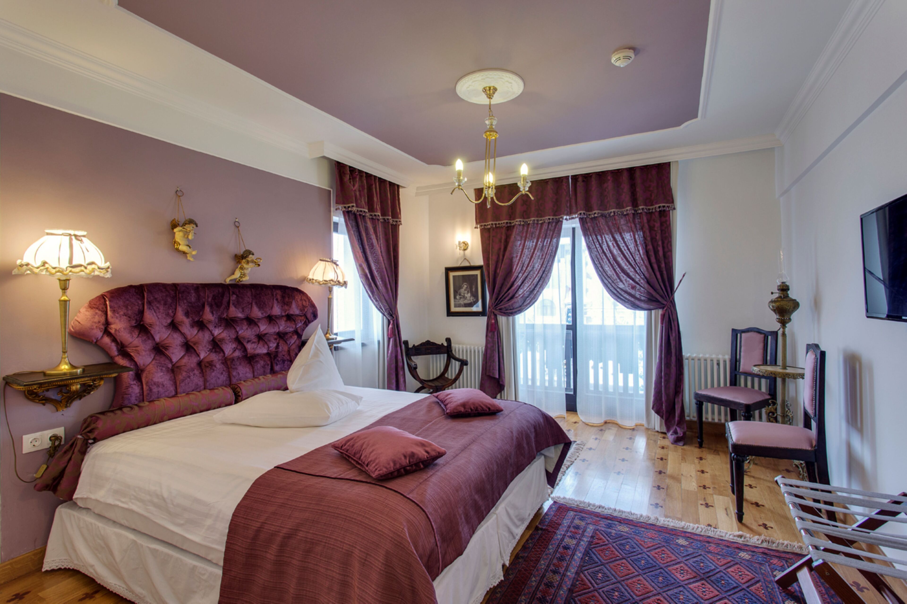 Ein Schlafzimmer mit einem großen Bett, einer Chandelüster, einem Schreibtisch mit Stuhl und Blick ins Freie.