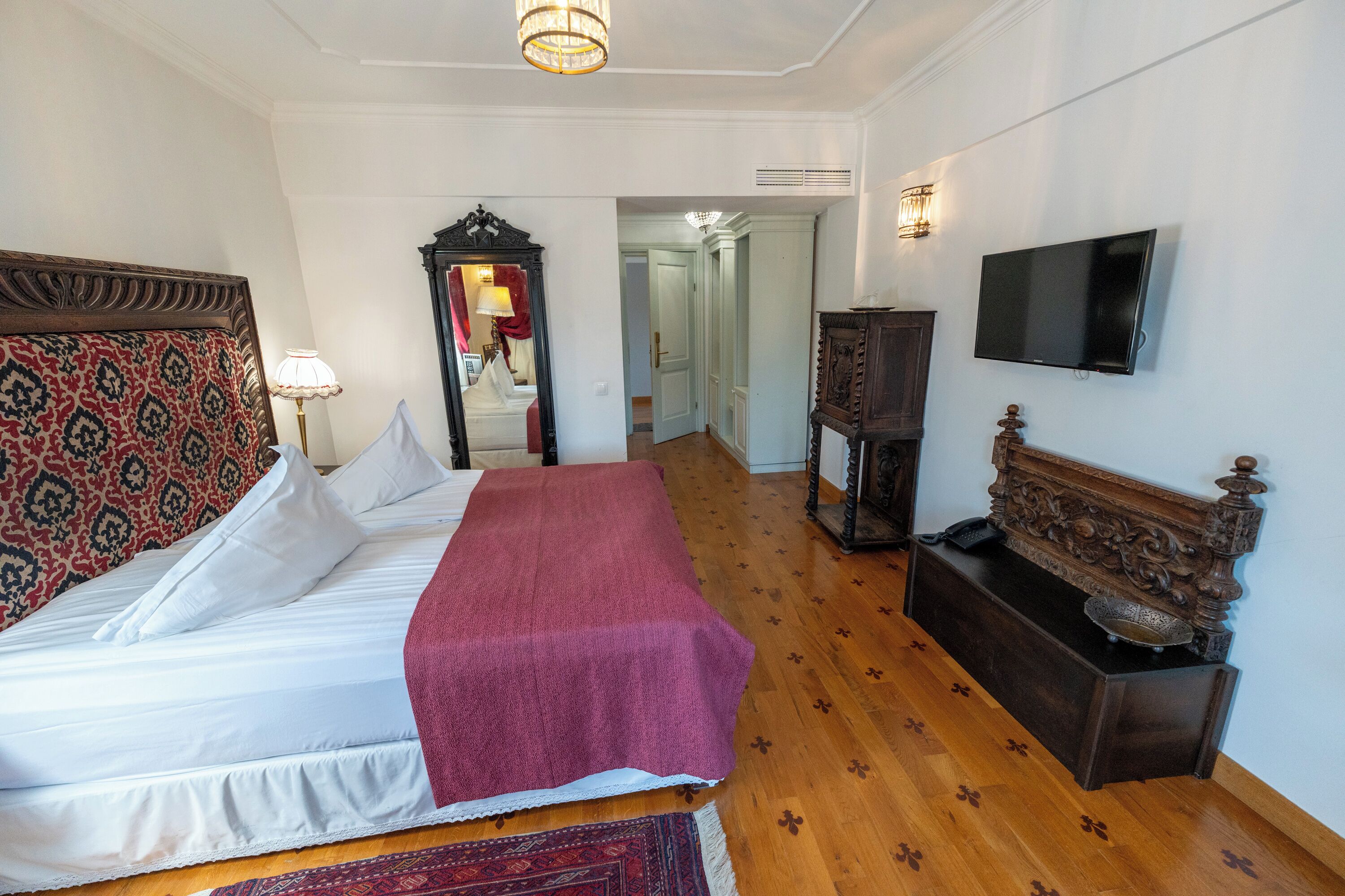 A hotel room with a large bed, a wooden floor, a TV mounted on the wall, and a mirror with a red curtain.