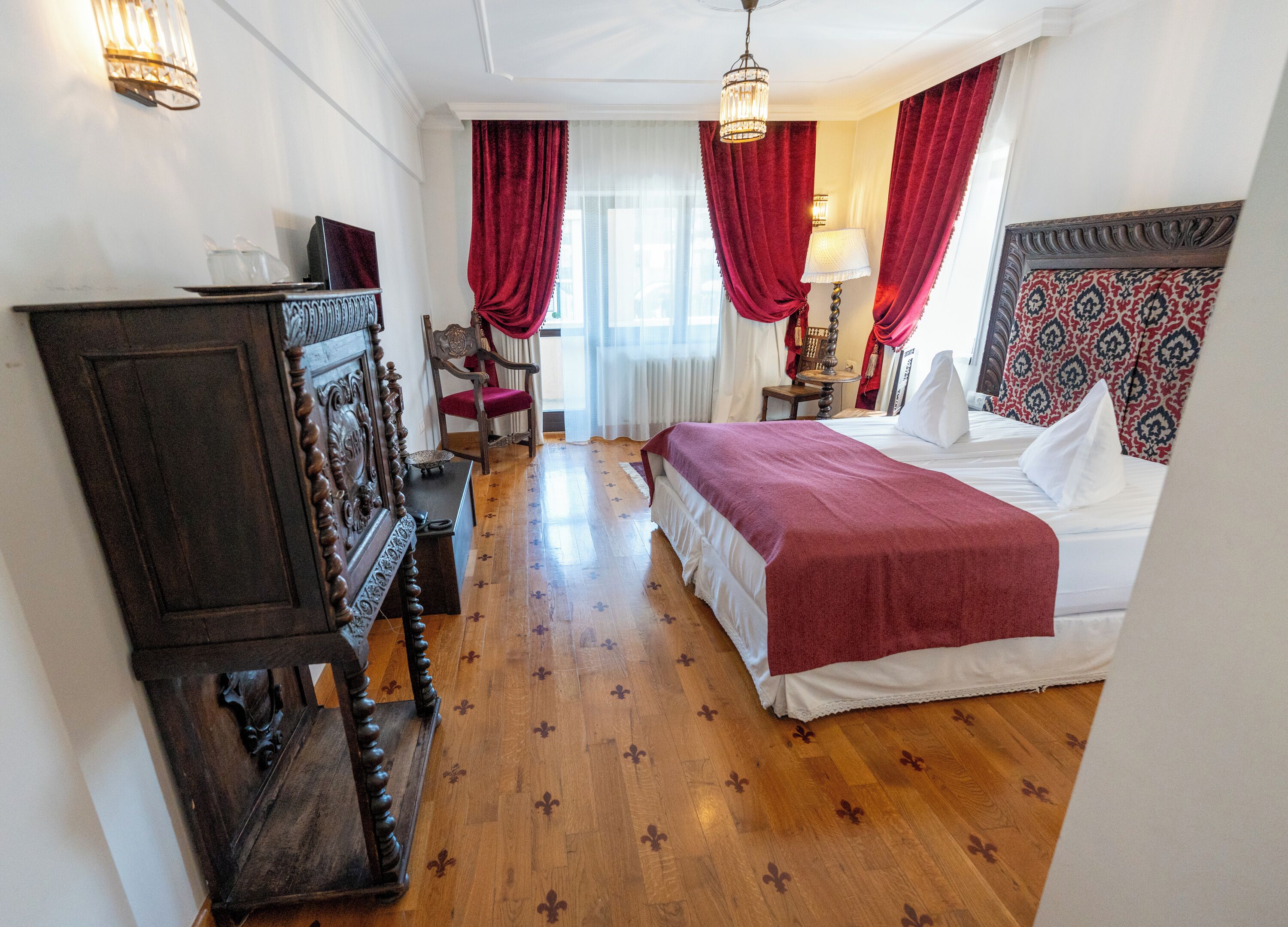 A hotel room with a wooden floor, a bed with white linens, a red bedspread, a wooden dresser, a chair, a lamp, and a patterned headboard.