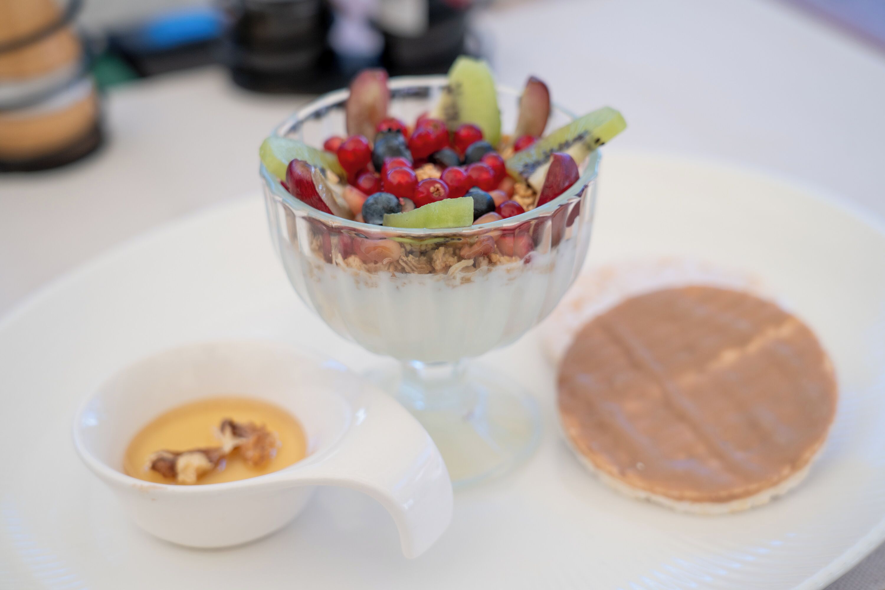 A dessert with a glass bowl containing yogurt, granola, and berries, a small bowl of sauce, and a pancake on a plate.