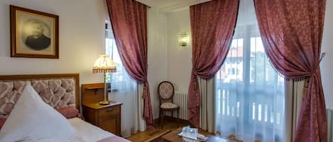 A hotel room with a bed, a chair, a table, a lamp, and curtains.