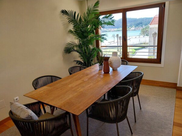 A dining area with a wooden table, black wicker chairs, a potted plant, and a view of a lake through a large window.