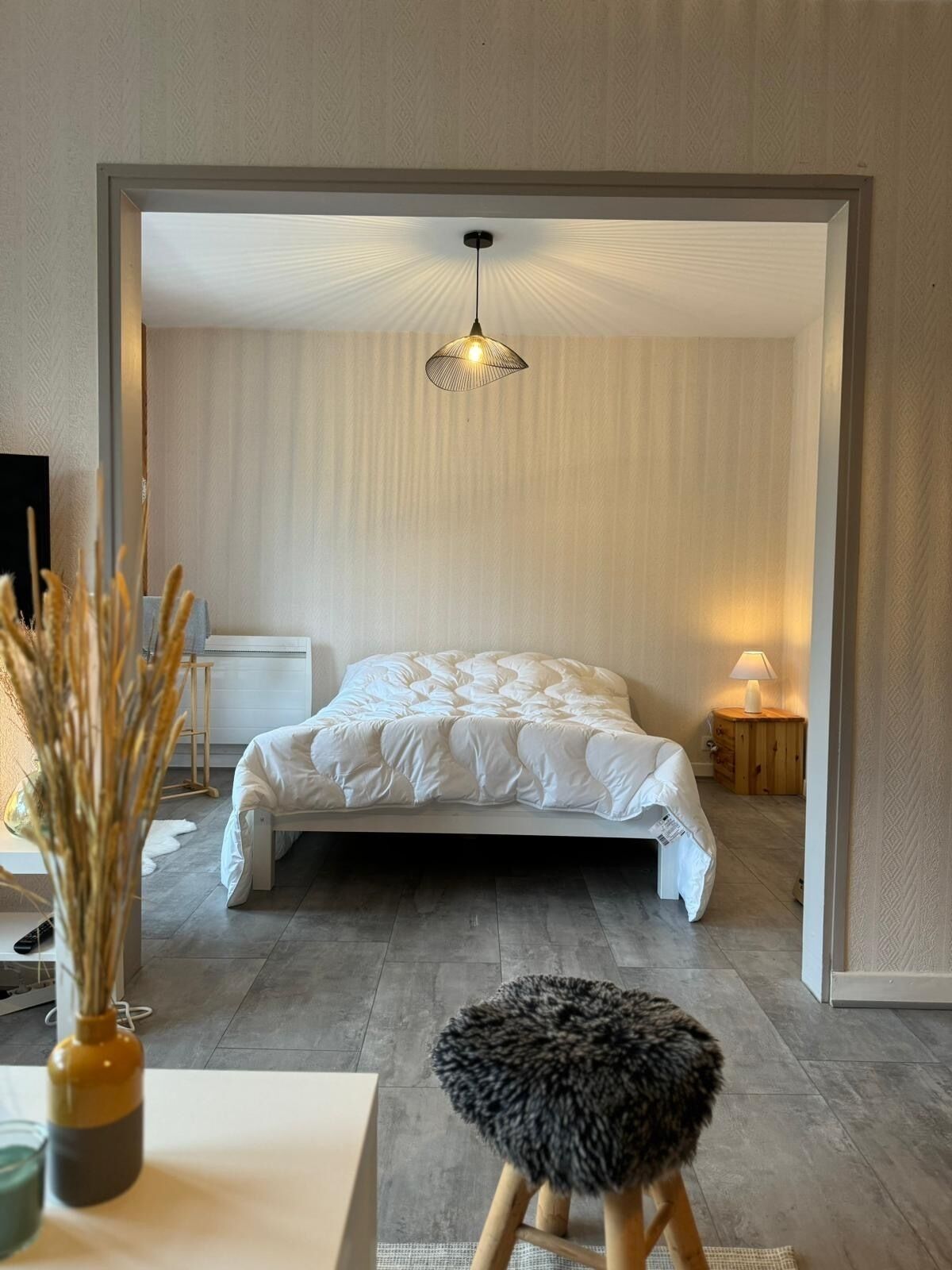 A bedroom with a bed, a hanging light fixture, a small stool, and a nightstand with a lamp.