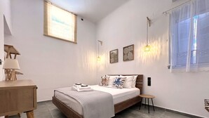 A modern bedroom with a wooden bed, a small bedside table, a desk with a lamp, and a window with sheer curtains. - Cycladic Home with a view  - 2 min walk from the beach!  (Adamantas)