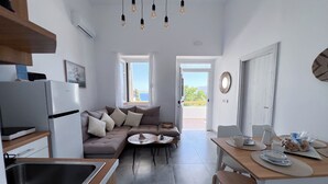 A modern kitchen with a built-in oven, a dining area with a table and chairs, a sofa with cushions, and a view through the open door. - Cycladic Home with a view  - 2 min walk from the beach!  (Adamantas)