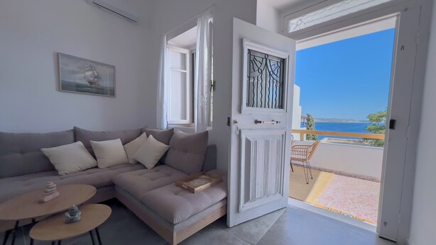 A living room with a grey sofa, a small round table, and a view of the sea through an open door. - Cycladic Home with a view  - 2 min walk from the beach!  (Adamantas)