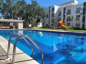 Una piscina con escaleras de metal, rodeada por un edificio con balcones y un parque infantil.