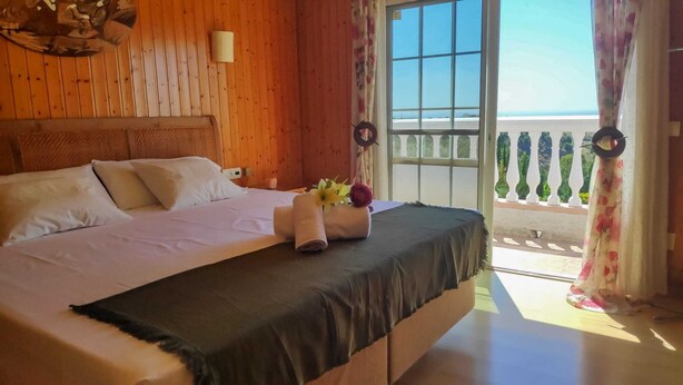 Ein Hotelzimmer mit einem hölzernen Kopfteil, einem Bett mit weißer Bettwäsche und Blick auf das Meer durch einen Balkon sowie einem Fenster mit blumigem Vorhang.