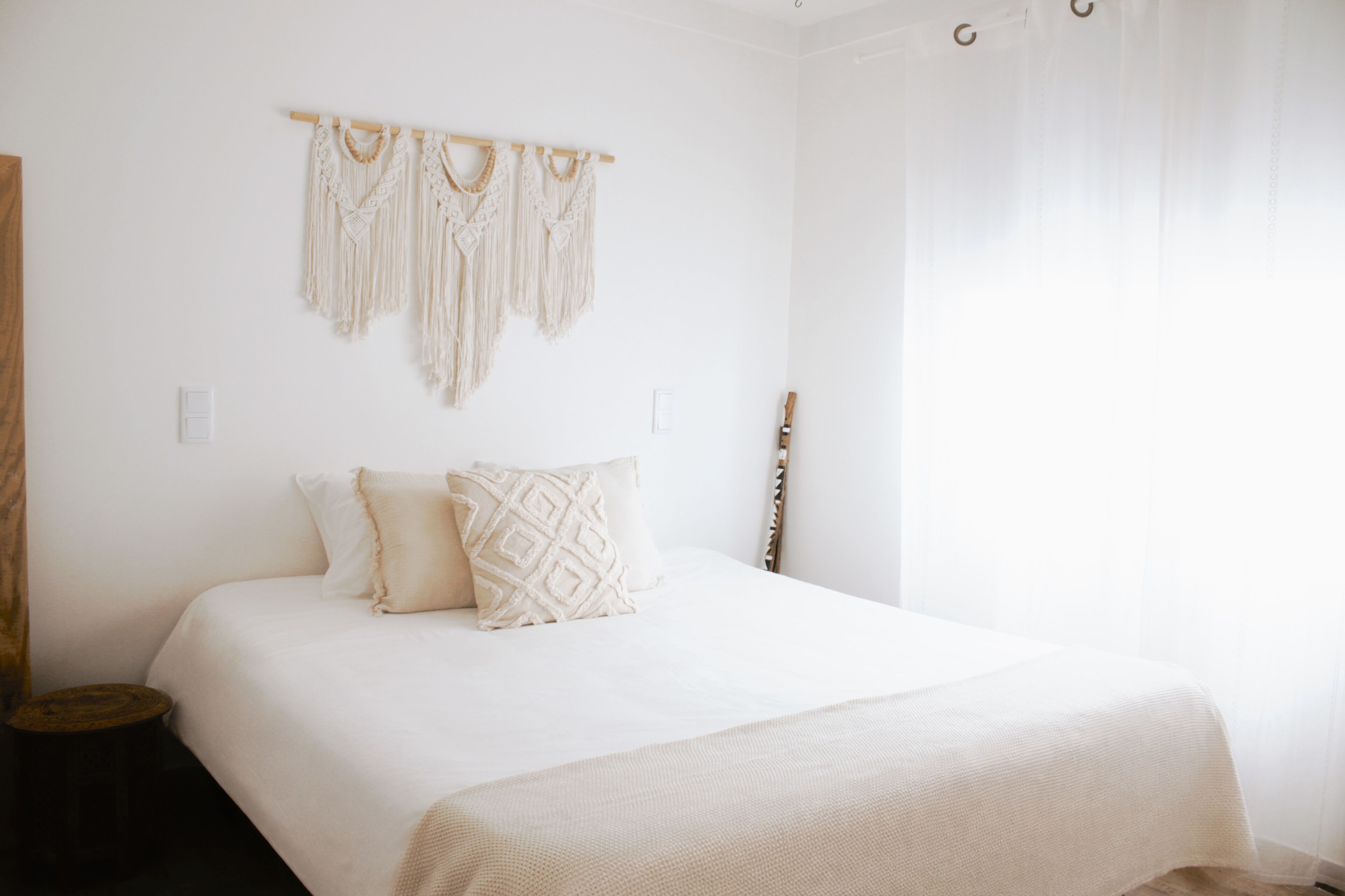 Een bed met een witte spreidek en twee gekleurde kussens, macramé decoratie aan de muur, en een houten hoofdbord.
