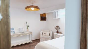 A bedroom with a bed, a chair with a 'HOLA' pillow, a bedside table, a wall-mounted shelf, and a hanging light fixture.