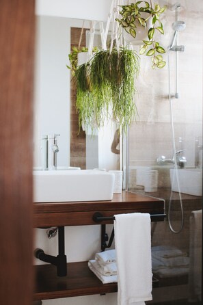 Un baño con tocador de madera, lavamanos blanco, espejo, planta verde colgante, toallero y zona de ducha.