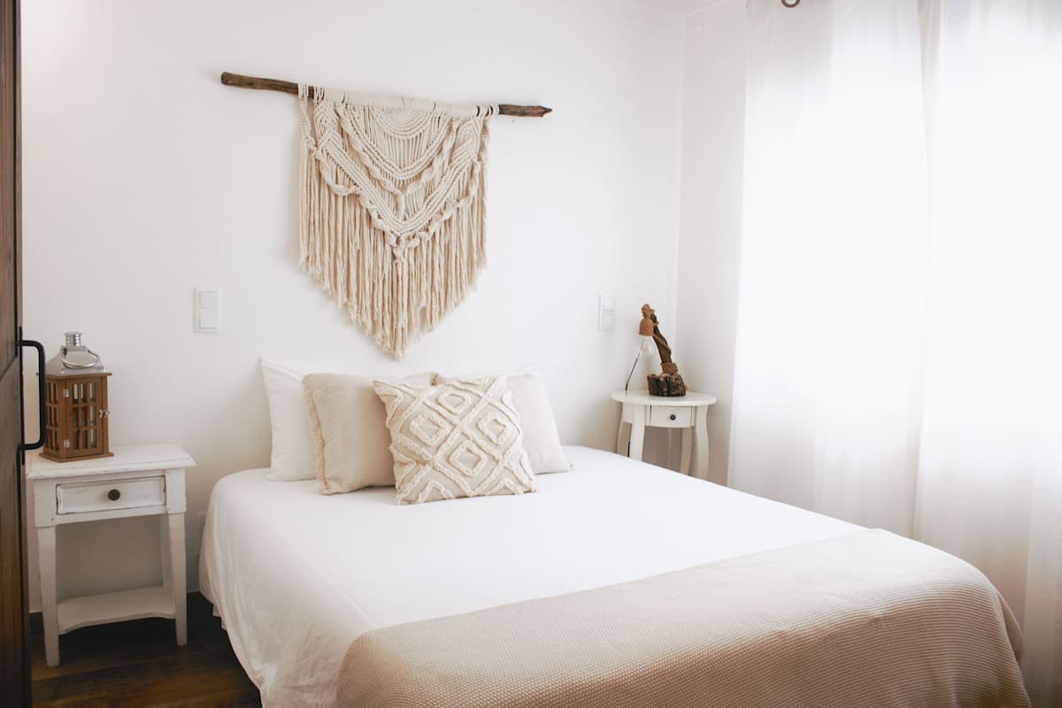 Un dormitorio con una cama, una mesita de noche, un colgador de macramé en la pared y una pequeña mesa con una lámpara.