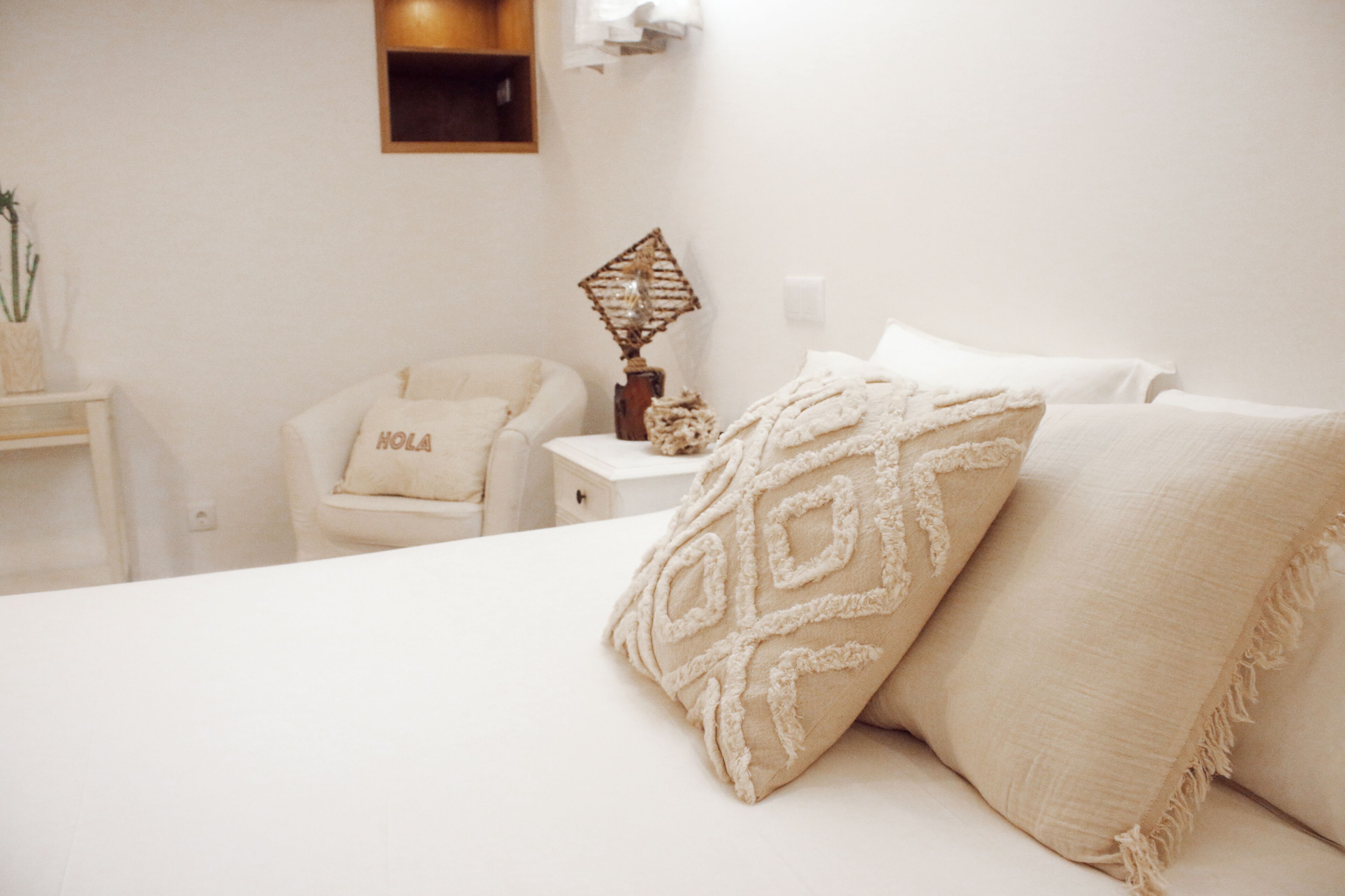 Een slaapkamer met een bed, kussens, een nachtkastje, een stoel met een 'HOLA' kussen, een wandplank en een wandlamp.