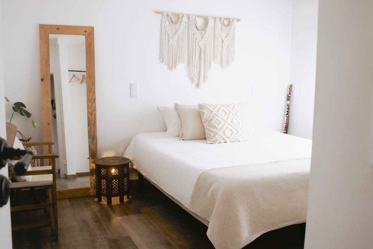 Un dormitorio con una cama, una silla de madera, una mesita redonda y un tapiz de macramé en la pared.