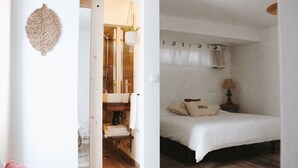 A bedroom with a bed, a bedside table, a hanging plant, and a bathroom visible through an open door. - Vila Vale Guest House surf e yoga (Almada)
