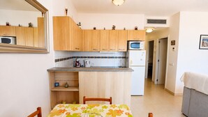 A compact kitchen with wooden cabinets, a microwave, and a refrigerator. There is a dining table with chairs and a floral tablecloth. - 224-Apartment with view on marina , pool and parking (Empuriabrava)