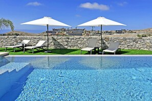 Piscina com água cristalina, rodeada por espreguiçadeiras e guarda-sóis, com uma parede de pedra e edifícios ao fundo.