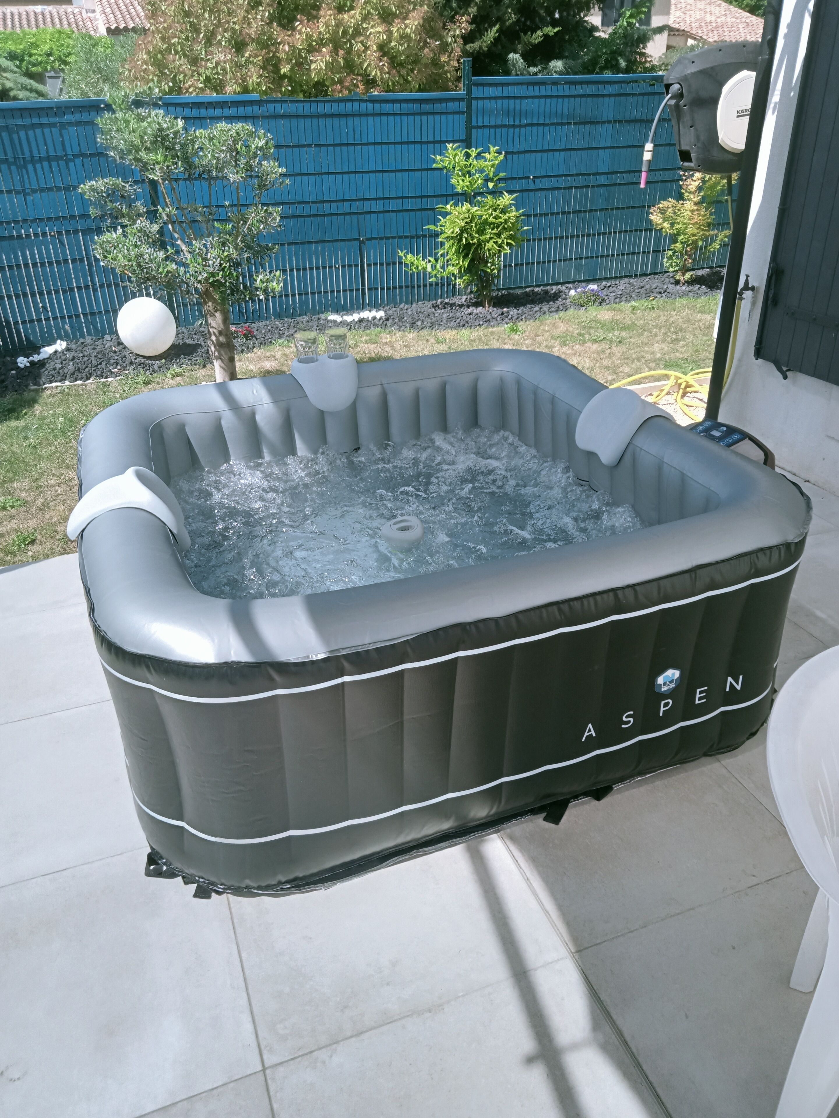 An inflatable hot tub with a control panel, placed on a patio with a blue fence and small trees in the background.