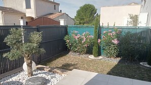 Ein Wohngarten mit einer gefliesten Terrasse, einem schwarzen Zaun und verschiedenen Pflanzen, darunter ein kleiner Baum und blühende Sträucher.
