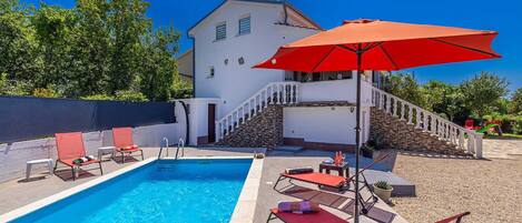 A swimming pool with lounge chairs and a red umbrella, surrounded by a white building with a stone staircase.