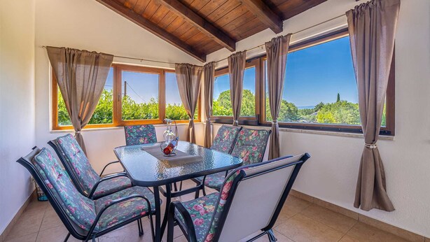 A dining area with a glass-top table and floral-patterned chairs, large windows with curtains, and a wooden ceiling. - Holiday house CHARMING with private pool (Kras)