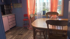 A kitchen with a microwave, a wooden table, and chairs. There is a window with curtains and a view of greenery outside.