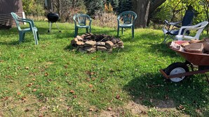 A grassy outdoor area with a pile of firewood, several plastic chairs, and a wheelbarrow.