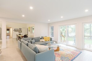 Una sala de estar moderna con un sofá modular, una mesa de centro de vidrio y una alfombra colorida. En el fondo, hay un comedor con una mesa y sillas.