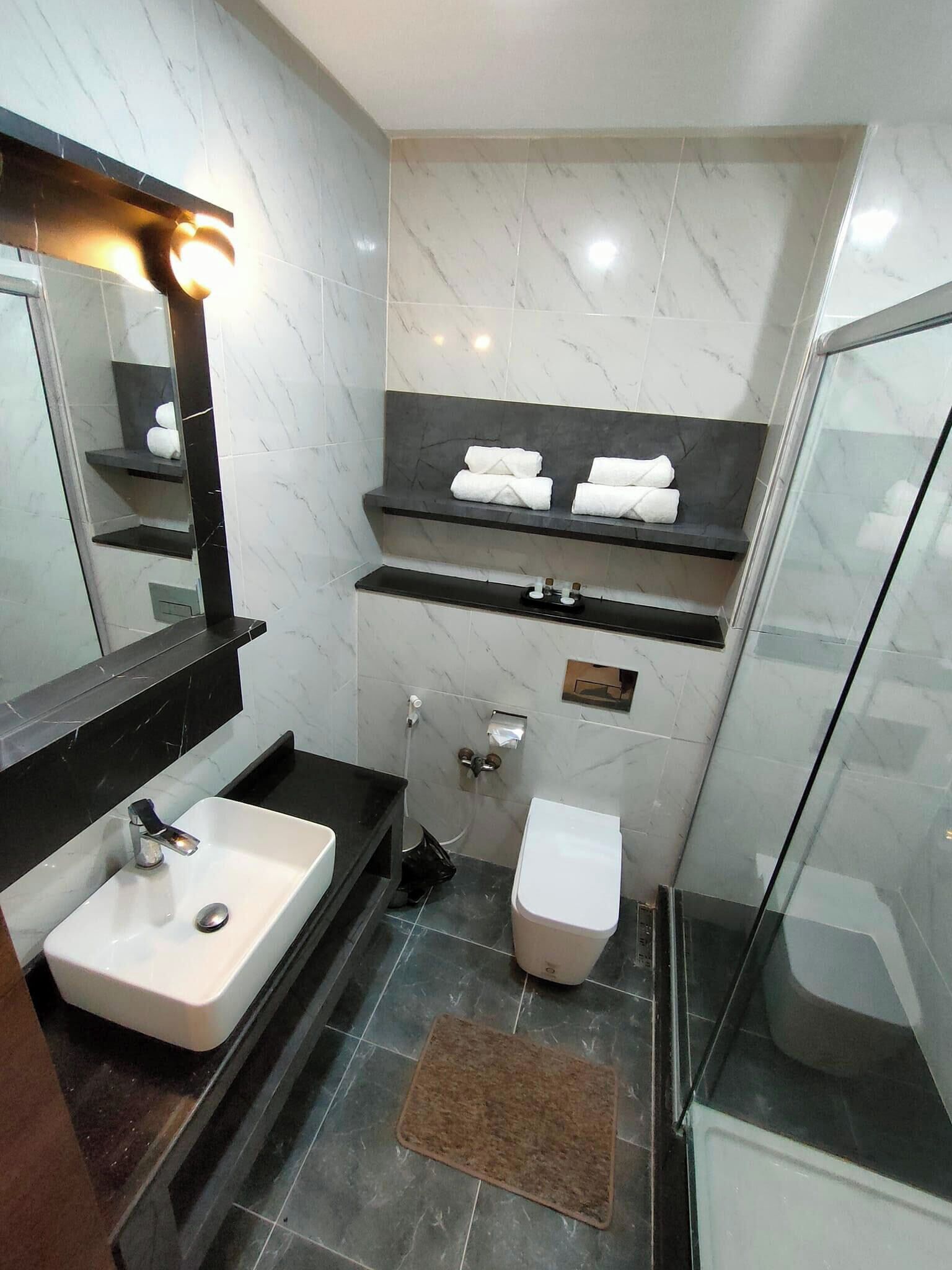 A modern bathroom with a white sink, dark countertop, and marble walls. There are two towel racks with towels, a glass shower enclosure, and a small rug on the floor.