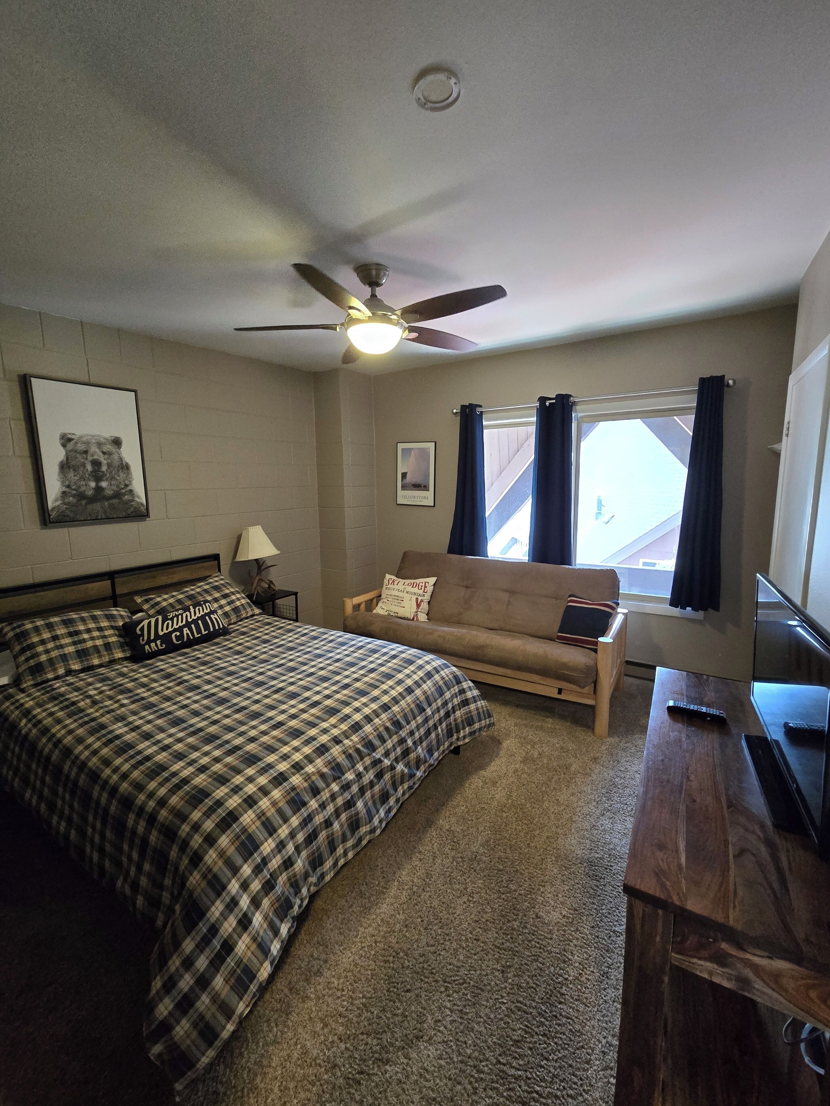A bedroom with a bed, a sofa, a ceiling fan, a TV, and a window with curtains.