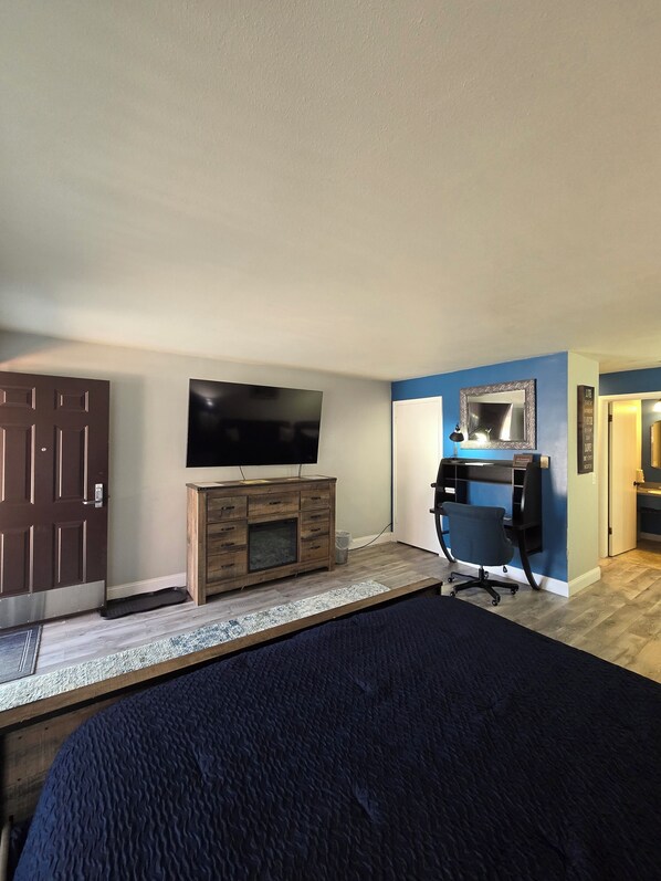 A bedroom with a bed, a TV, a wooden dresser, a desk with a chair, and a door.